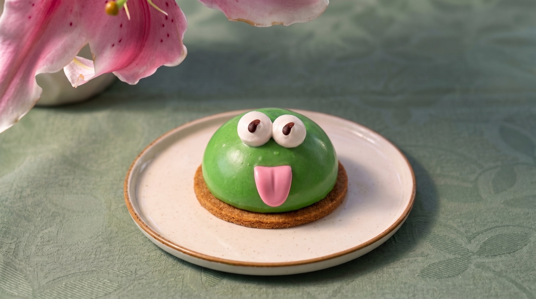 Ranita de Friandise: pastelito verde con ojos y lengua rosada sobre base de galleta, junto a una flor de lis rosada